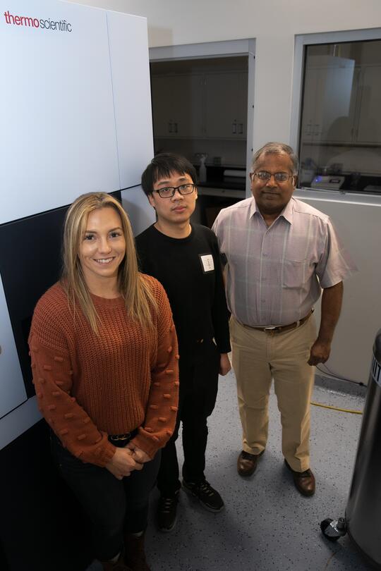 Left to right: Drs. Kristen Flatt, Yiwu Zheng, and Satish Nair stand together in front of a cryogenic electron microscope. They are all smiling for the camera.